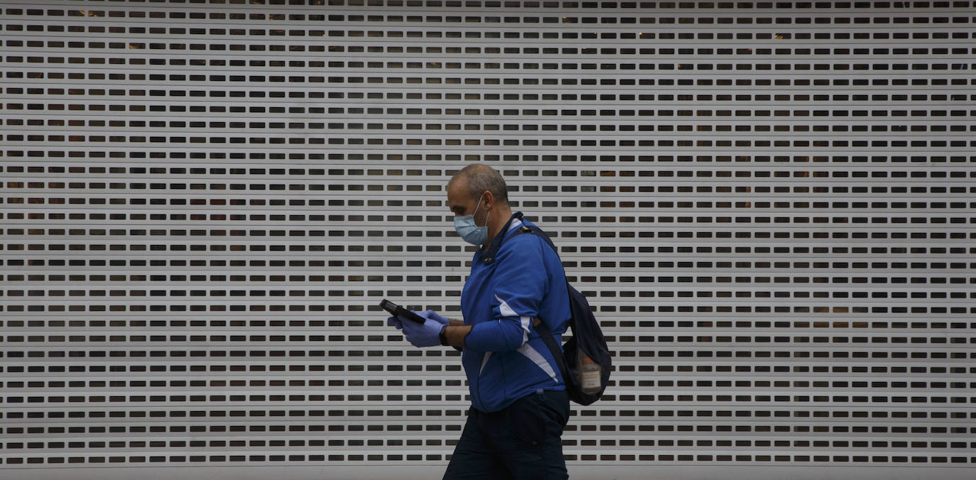 Un hombre con guantes y mascarilla pasa junto a la verja cerrada de un comercio. 