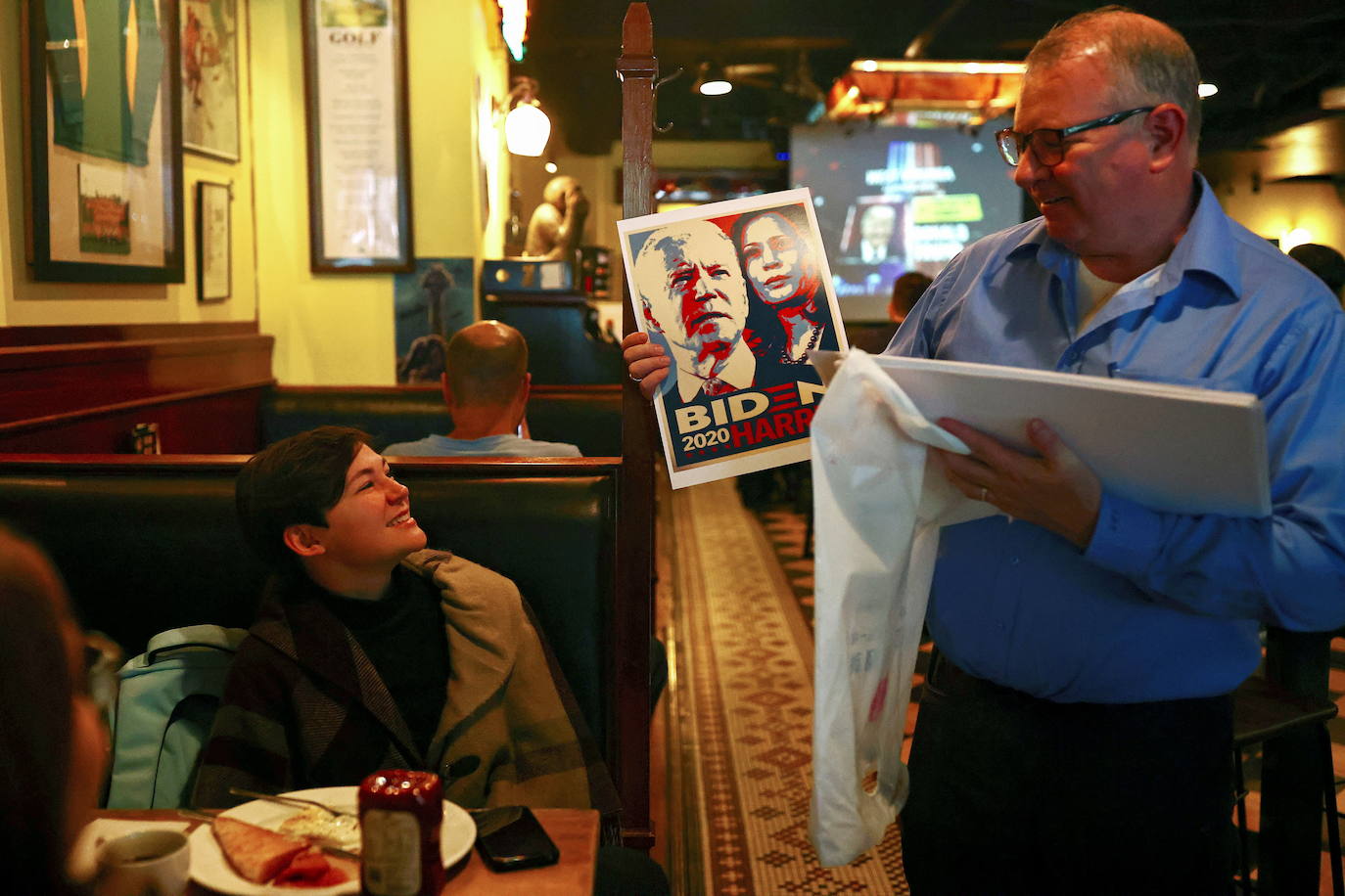 Un hombre sostiene un cartel con la imagen de Biden y la senadora Kamala Harris mientras otra gente ve los resultados en vivo de las elecciones. 