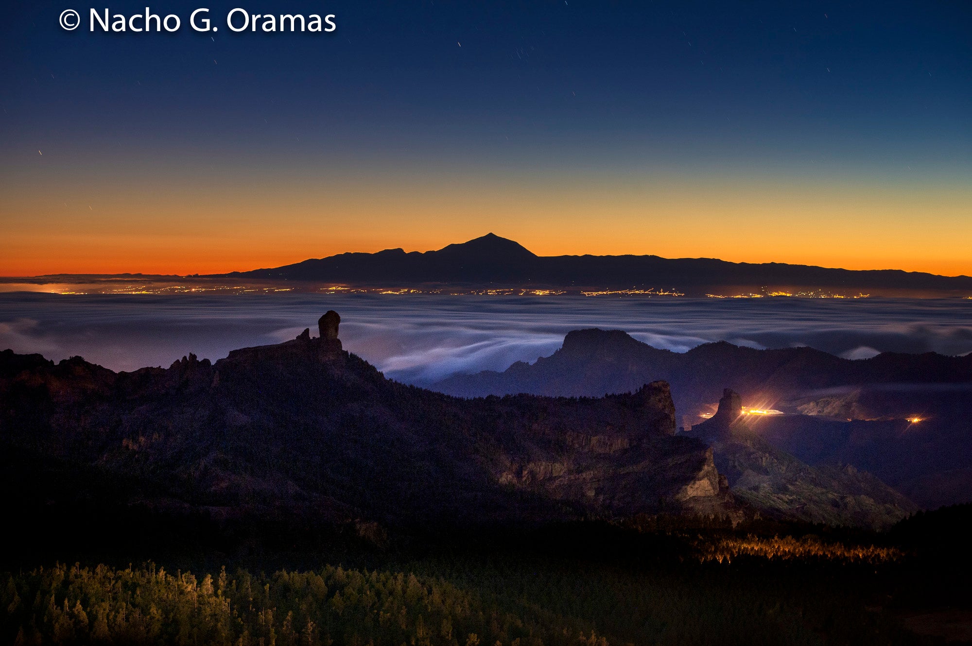 El pico Teide y las luces de la costa este de Tenerife sobre el Roque Nublo, La Fogalera, el Roque Bentayga y la montaña de Altavista, en Tamadaba (Pico de Las Nieves, Tejeda).