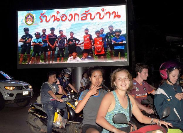 Jóvenes en moto celebran la buena noticia mientras pasan ante un cartel con la imagen de los jóvenes rescatados. :: TANG CHHIN Sothy / afp