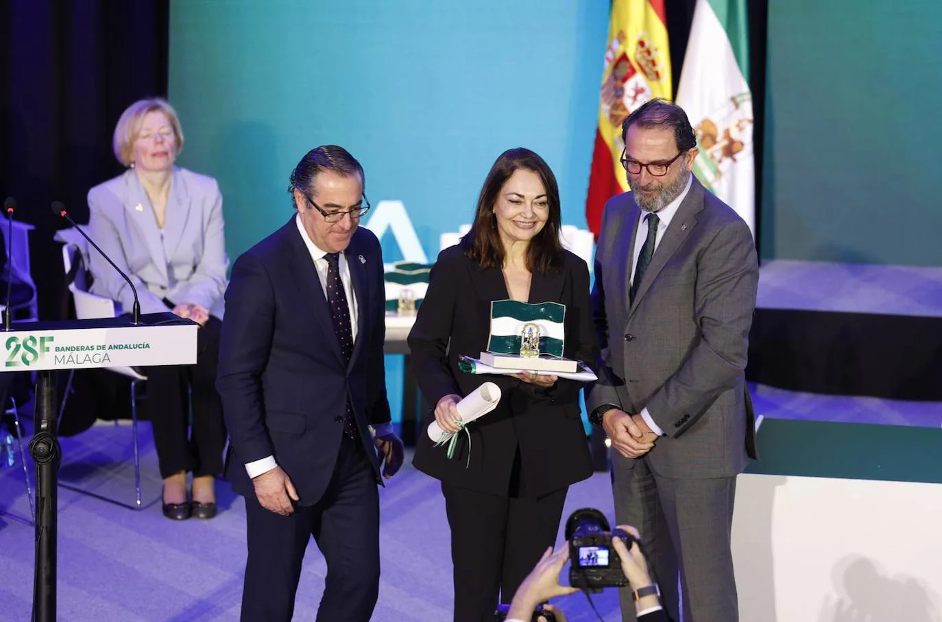 La catedrática de la UMA Gloria Corpas recibe la Bandera de la Investigación, la Ciencia y la Salud. Le entregan el premio los delegados Carlos Bautista y Miguel Briones
