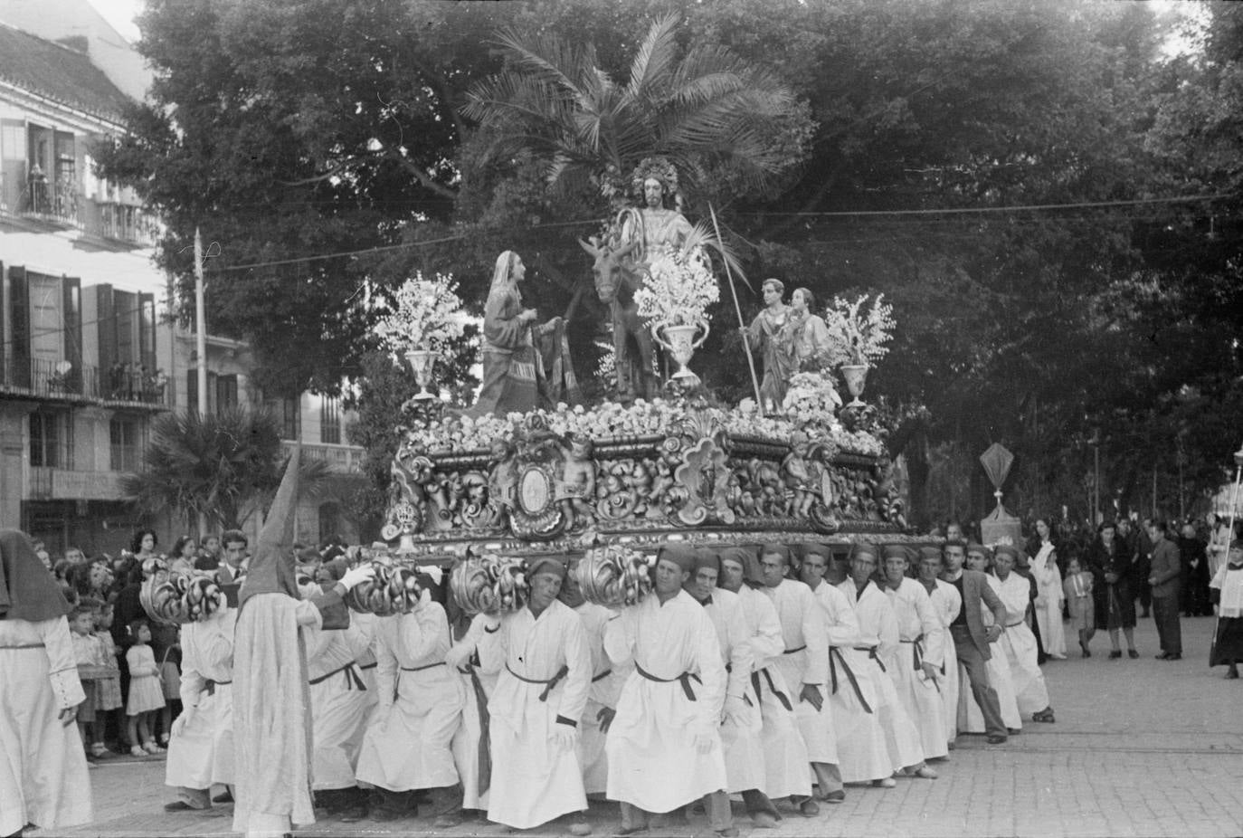 Los hombres de trono meten el hombro para levantar el conjunto procesional del Cristo de la Cofradía de la Pollinica, que se dispone a abandonar la Alameda Principal para embocar la rotonda del Marqués de Larios. El trono es el realizado por Antonio Castillo Ariza y Francisco Díaz en 1945, sustituido por el actual, que se estrenó el Domingo de Ramos de 2015.