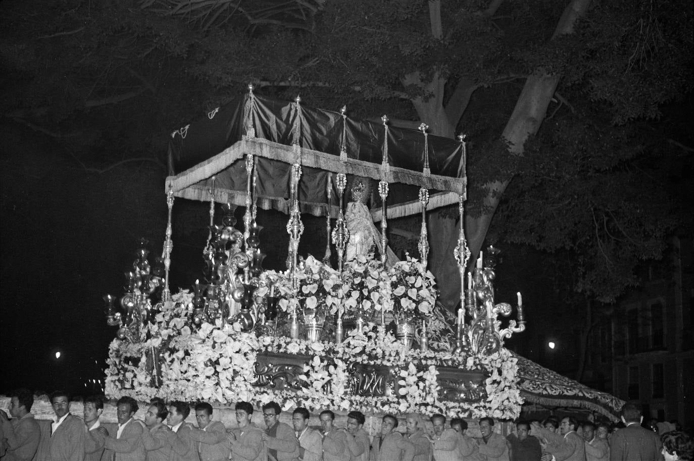 La Virgen de la Concepción bajo un palio de cajón, sin bordados, en la noche del Domingo de Ramos de 1959. En la instantánea se aprecia el soberbio manto de la Dolorosa, ejecutado en los talleres de Leopoldo Padilla en 1947, que presenta un enorme olivo con esmeraldas prendidas y una magnífica escena, bordada en sedas, de la Oración en el Huerto.