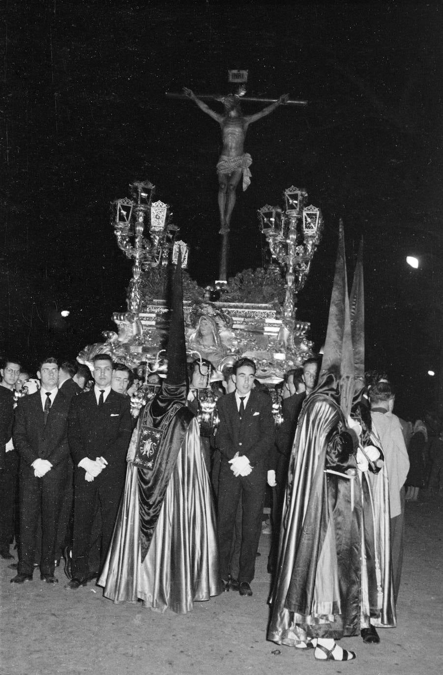 En esta fotografía, correspondiente al Martes Santo de 1959, se aprecia el antiguo Cristo de la Agonía, obra de Pedro Pérez Hidalgo, tallado en 1948. El Crucificado figura sobre un trono que fue ejecutado por el propio Pérez Hidalgo en 1949, caracterizado por sus cuatro arbotantes de madera rematados con faroles. Las andas procesionales sirvieron, asimismo, para portar la imagen actual, de Francisco Buiza, hasta que se estrenó el trono de Antonio Martín en 1982.