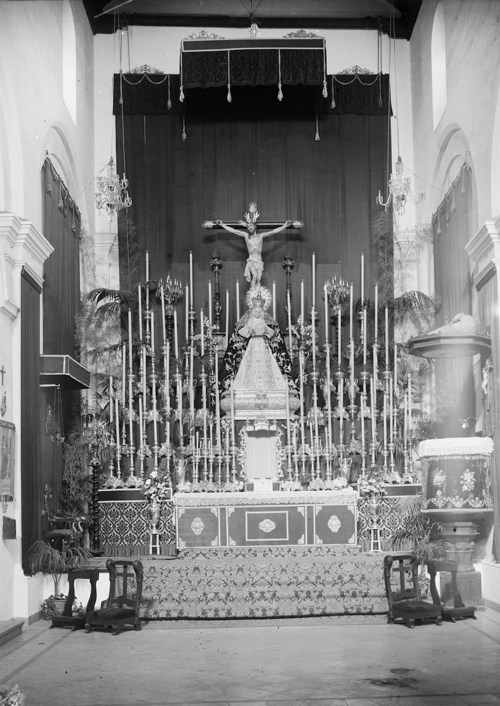 En los años cincuenta, la Archicofradía de la Expiración montaba en el presbiterio de San Pedro enormes altares de cultos como el que aparece en la fotografía, en la que el Cristo estaba situado muy alto, y la Virgen de los Dolores, a sus pies. Pocas cofradías podían llevar a cabo tales montajes, pero el patrimonio de la Expiración sí lo permitía. La candelería que aparece en la imagen es la de cultos, con velas que culminan en una bombilla. Toda una odisea de altar efímero para la época en la que tenía mucho que ver la labor de Rafael Alfonso García-Hidalgo, que durante muchos años fue el vestidor de la Virgen de los Dolores. Según apuntan desde la archicofradía, en este caso en concreto, el altar se instaló con motivo de la proclamación del dogma mariano de la Asunción, el 1 de noviembre de 1950.