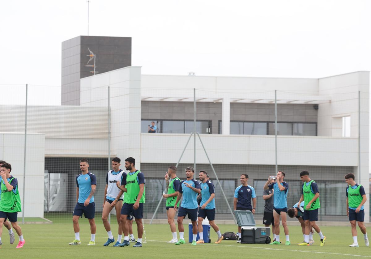 Jugadores del Málaga se refrescan durante el entrenamiento de este lunes en La Academia, con el edificio principal de la ciudad deportiva de fondo.