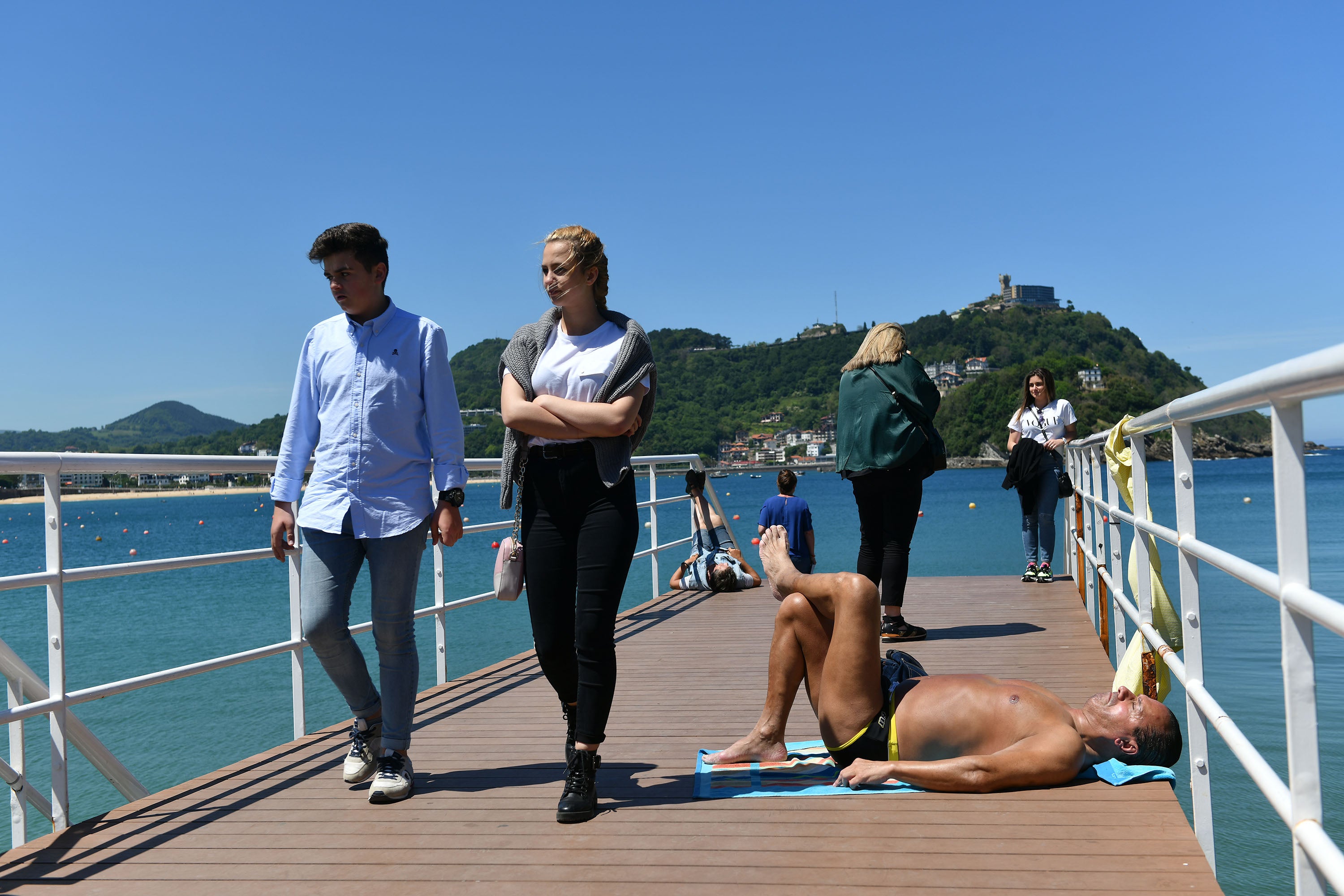 Un hombre toma el sol en la pasarela del Náutico en Donostia mientras la gente pasea. 