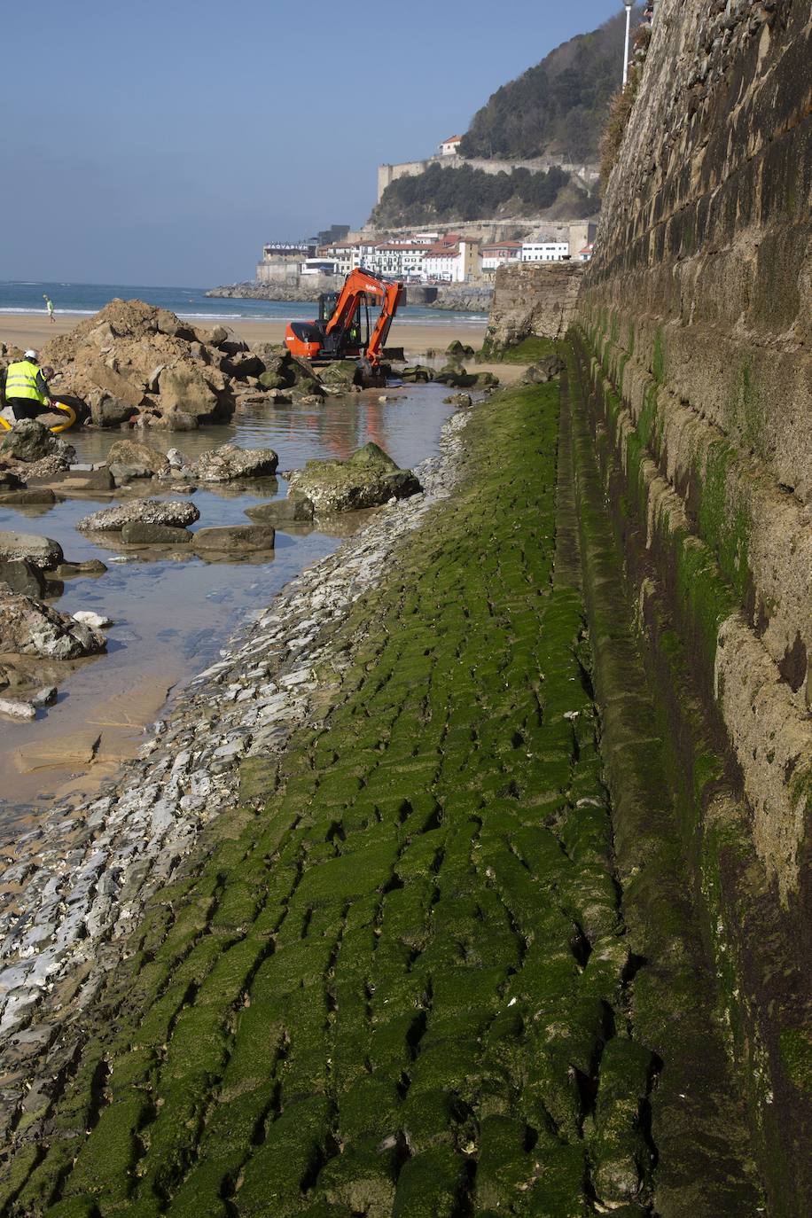 La playa de La Concha se ha convertido en la bajamar de este lunes por la mañana en un inesperado yacimiento arqueológico. La presencia de una pequeña excavadora retirando piedras y escombros junto al muro de costa a la altura del edificio consistorial ha suscitado una gran expectación entre los numerosos viandantes y turistas que han provechado el buen tiempo para dar un paseo. La máquina profundizaba en la arena en busca de los restos de un barco hundido del que hay constancia documental desde el siglo XVIII. 