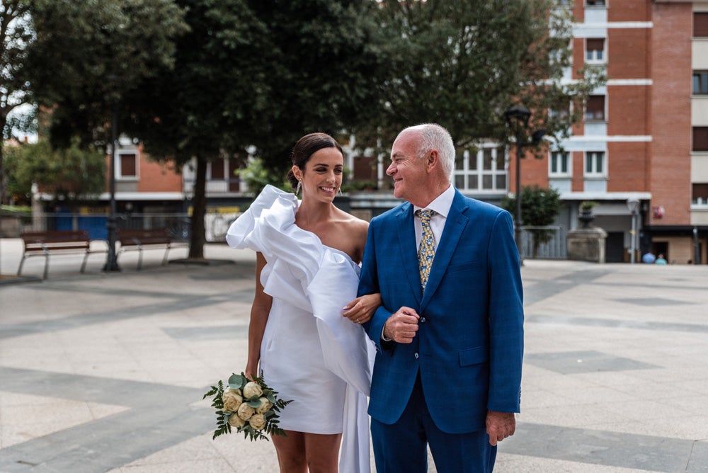 María y Jorge se casaron en Gorliz en una ceremonia íntima adaptada a las circunstancias. De largura 'mini' por encima de la rodilla, con corte asímetrico, volumen en la parte superior y lazadas laterales, la protagonista se ha convertido en una de las novias más impresionantes de las últimas semanas. El diseño lo ha firmado Fabio Encinar, uno de los diseñadores con sede en Madrid más de moda. En cuanto al peinado, otro de los elementos más destacados del estilismo, ha optado por un acertado moño bajo de bailarina súper pulido y tirante, con horquillas de Oui Novias