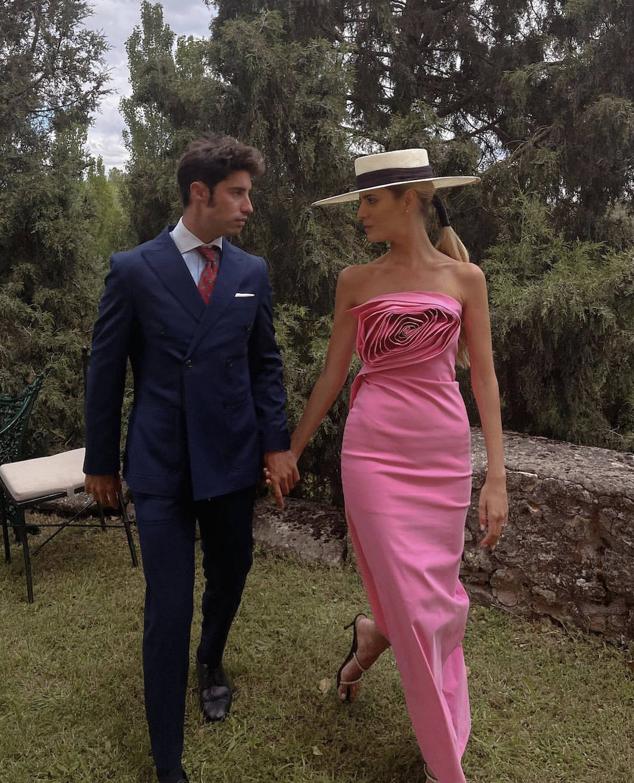 Teresa Andrés Gonzalvo, 'influencer' que protagonizará el próximo enlace mediático con Ignacio Ayllón, ha seleccionado un vestido tubo palabra de honor de largura 'midi', realizado en tafeta de seda rosa y decorado con una gran flor en la zona del pecho. El diseño lo ha firmado Juan Vidal y lo ha combinado con un sombrero de ala ancha con cinta negra de Mimoki y unas sandalias de tiras. El escote y la flor con volumen ha recordado al que ya lució en la boda de Marta Lozano hace un mes en Alicante. El maquillaje ha sido obra de Dior
