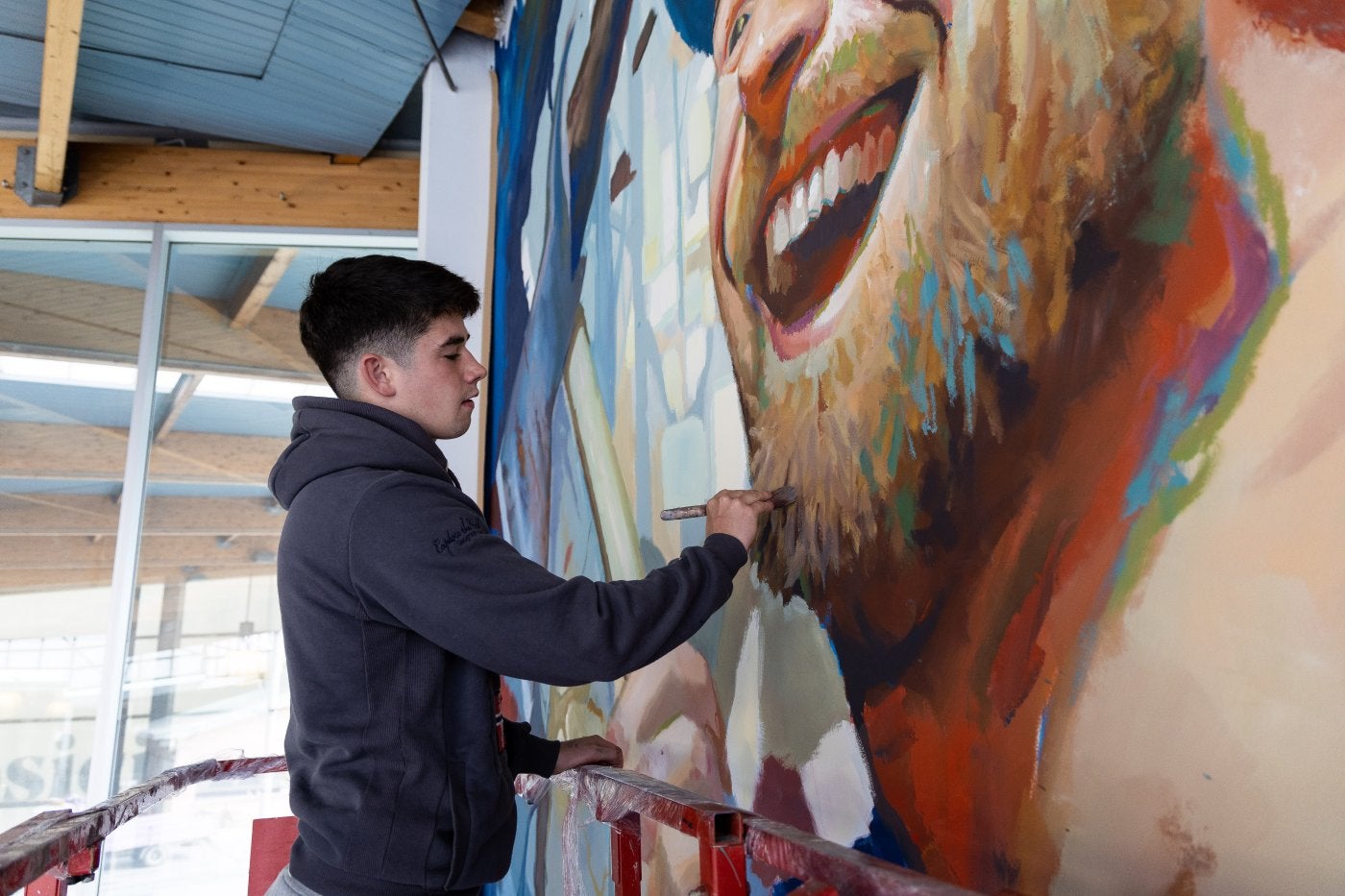 Imagen principal - Arriba, Nexgraff da los últimos retoques a uno de sus murales. En medio, la escuela pública dde Villabona cuenta con dos murales de Nexgraff. Debajo, Nexgraff con un bote de pintura azul.