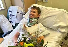 Jay Vine, en su habitación del hospital con un collarín y un ramo de flores, levanta el pulgar de su mano derecha y sonríe feliz de ver cómo avanza en su recuperación.