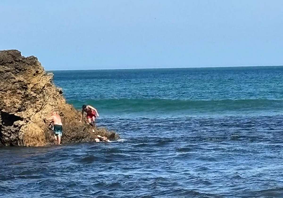 El hombre se agarra a la roca, mientras en el agua el ertzaintza sujeta a la mujer exhausta y el hombre del bañador rojo se acerca para ayudarlos.
