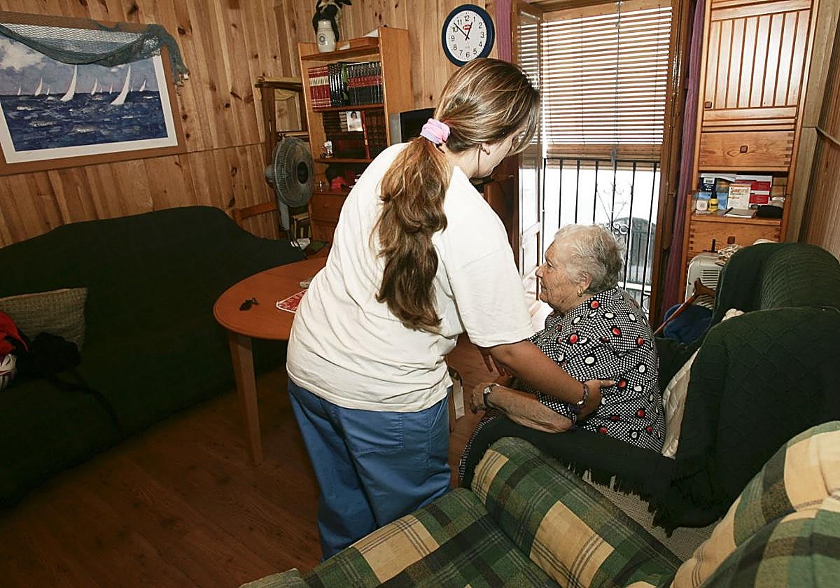 Una mujer es ayudada a levantarse del sillón por su cuidadora.