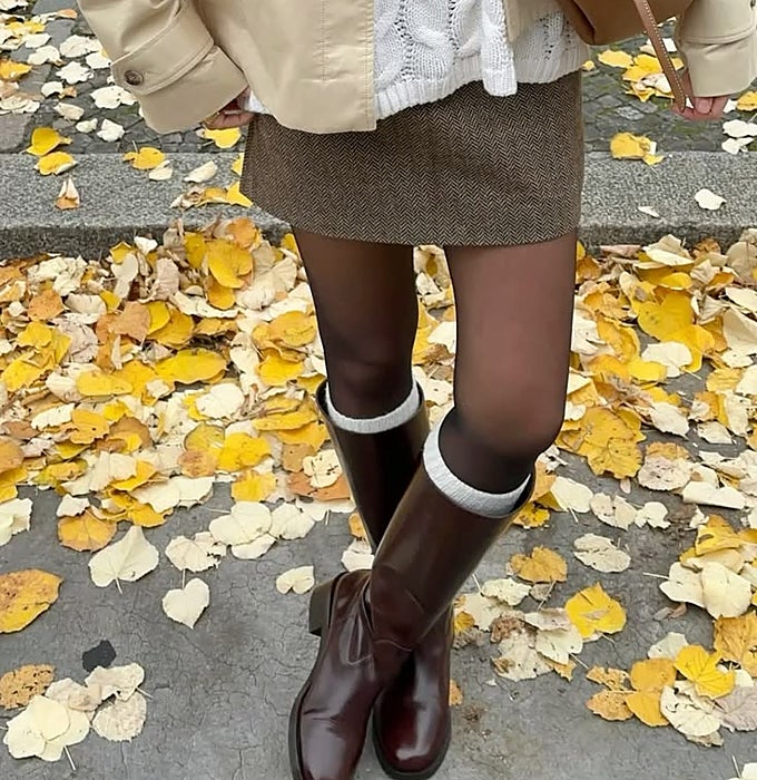 Cazadora beige, jersey blanco de punto trenzado y minifalda marrón, con botas altas de piel en granate y calcetines grises.
