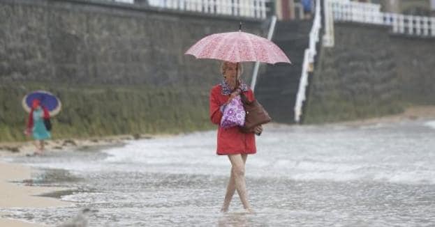 Una mujer, con paraguas y chubasquero, en la orilla de la gijonesa playa de San Lorenzo. 