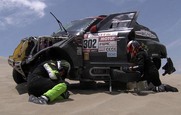 El copiloto Álex Haro trata de reparar el Mini, con Nani Roma en el suelo, sobre la arena. 