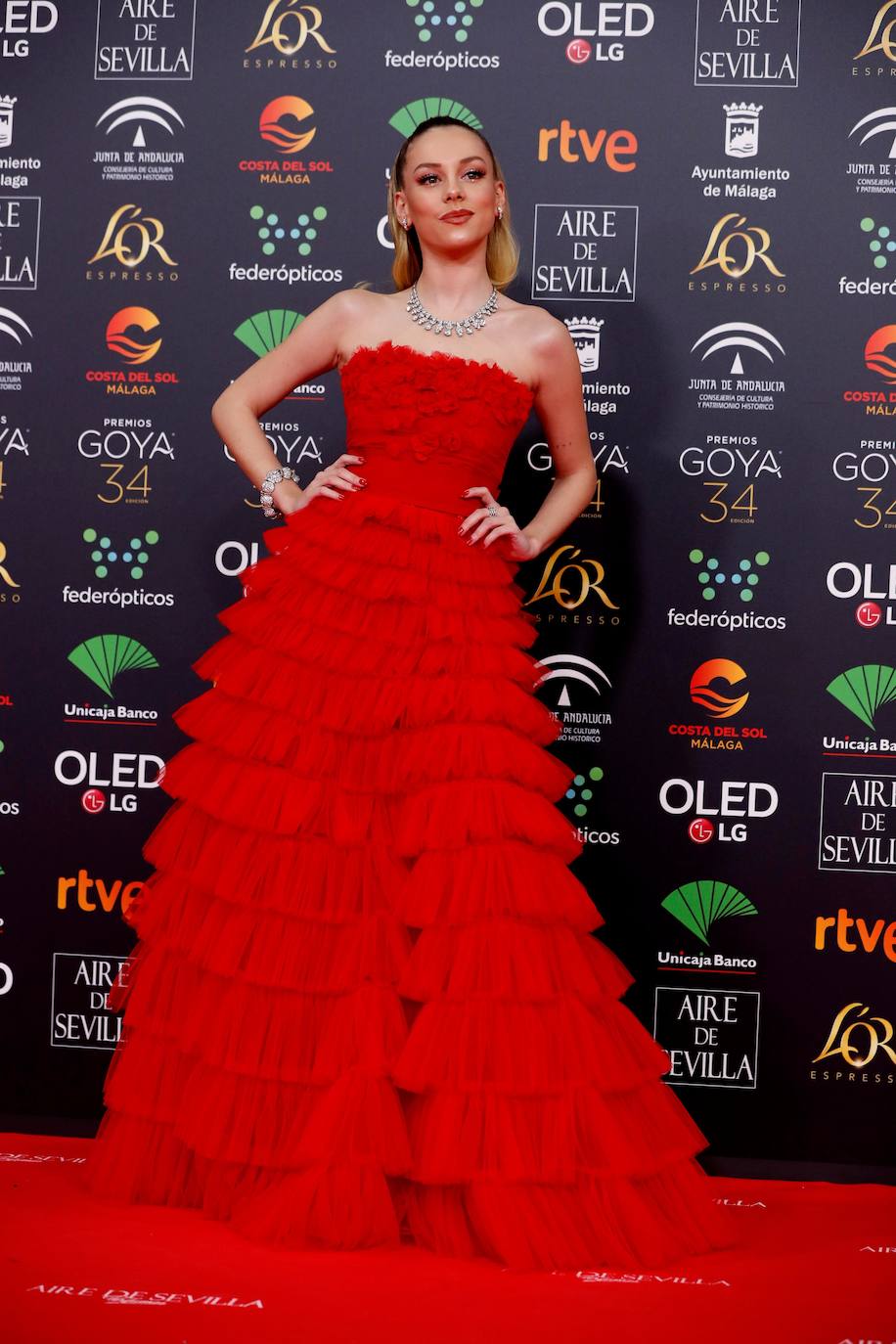 Ester Expósito añade la nota de color con un vestido rojo firmado por Twinset. Es un diseño con escote palabra de honor, cuerpo drapeado con flores en relieve y una falda con volumen confeccionada a base de microvolantes. El maquillaje, de Yves Saint Laurent, fue ideado por Gino Mateus y marcaba la mirada de la actriz con sombras en tonos tierra.