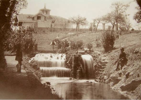 Estanque y cascada de La Noria con La Isla primitiva al fondo y dos operarios del servicio acondicionando el juego hidráulico. (Fecha: hacia 1896)