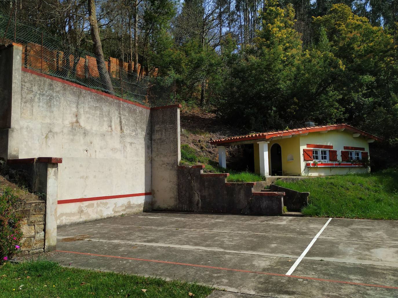 Frontón, cancha de tenis y vestuarios con el bosque mixto de pinos, robles y eucaliptos detrás.