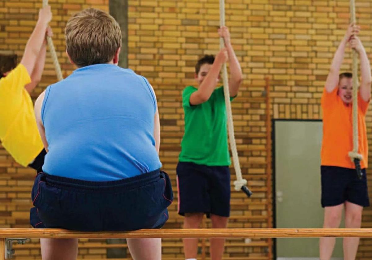 Un niño con sobrepeso observa a dos compañeros realizar ejercicios en el gimnasio de un un colegio.