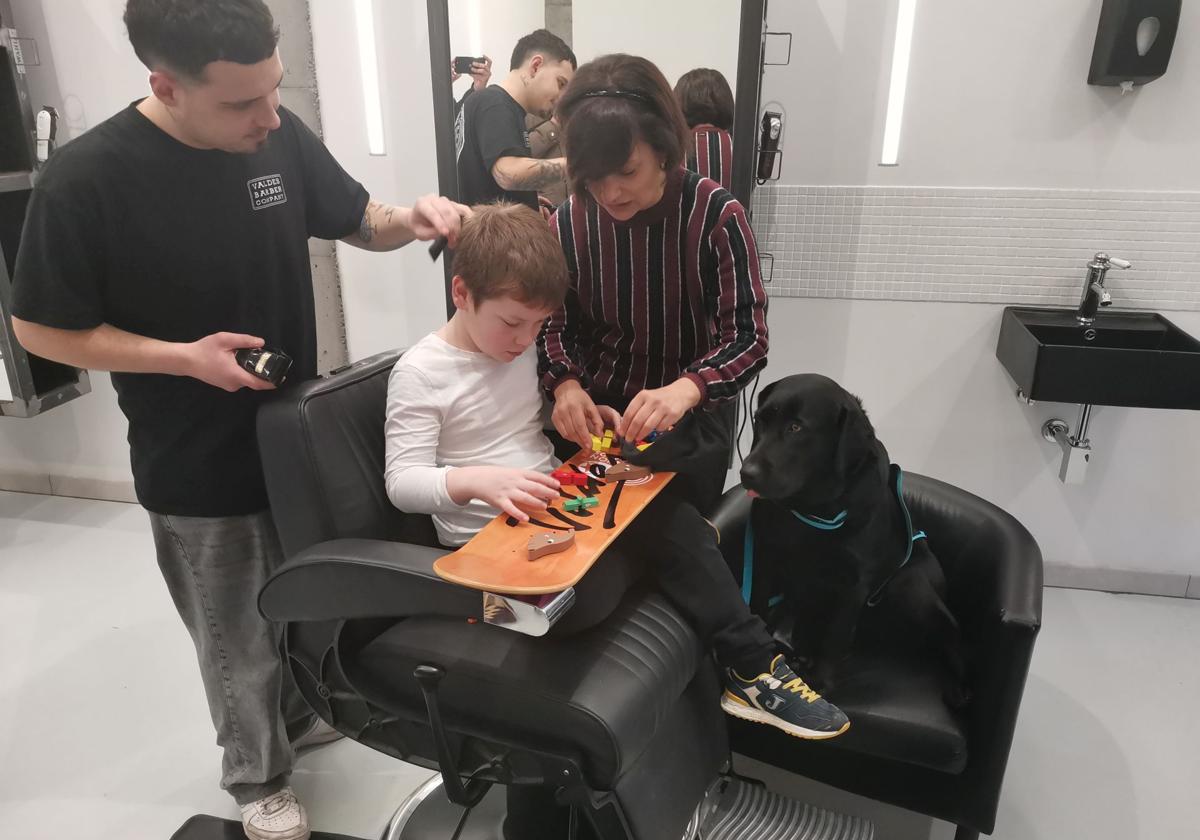 El pequeño Xuan López recibe el corte de pelo en la peluquería Valdés Barber Company, acompañado de la perra labrador 'Boni' y su adiestradora, Hortensia Vidal, de la Asociación Trufa Negra