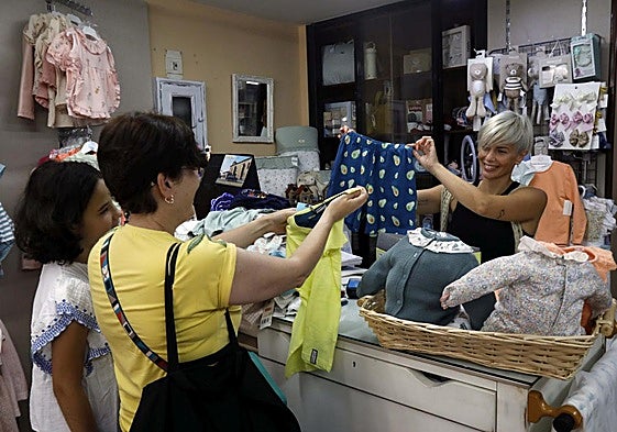 Leticia Rodríguez, de Leticia Mercería Lencería Bebé, muestra unos pantalones de niño a una clienta y a su hija.