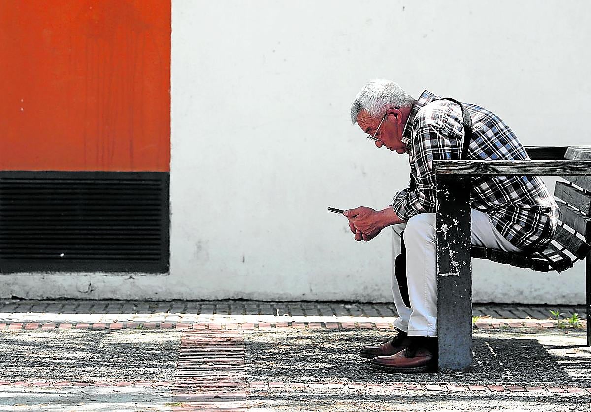Un jubilado observa su móvil sentado en un banco a la sombra.