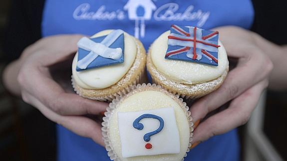 La empleada de una pastelería posa sujetando una magdalena decorada con un interrogante junto a otras dos con la bandera de Escocia y la del Reino Unido. 