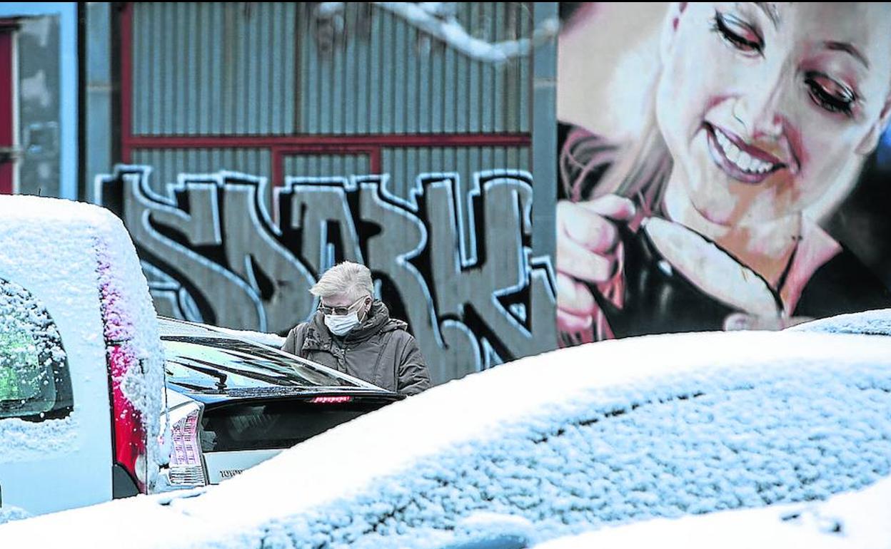 Sin bufanda pero con mascarilla, una señora camina entre los coches cubiertos de nieve ante un mural con una mujer bebiendo de una taza humeante. 