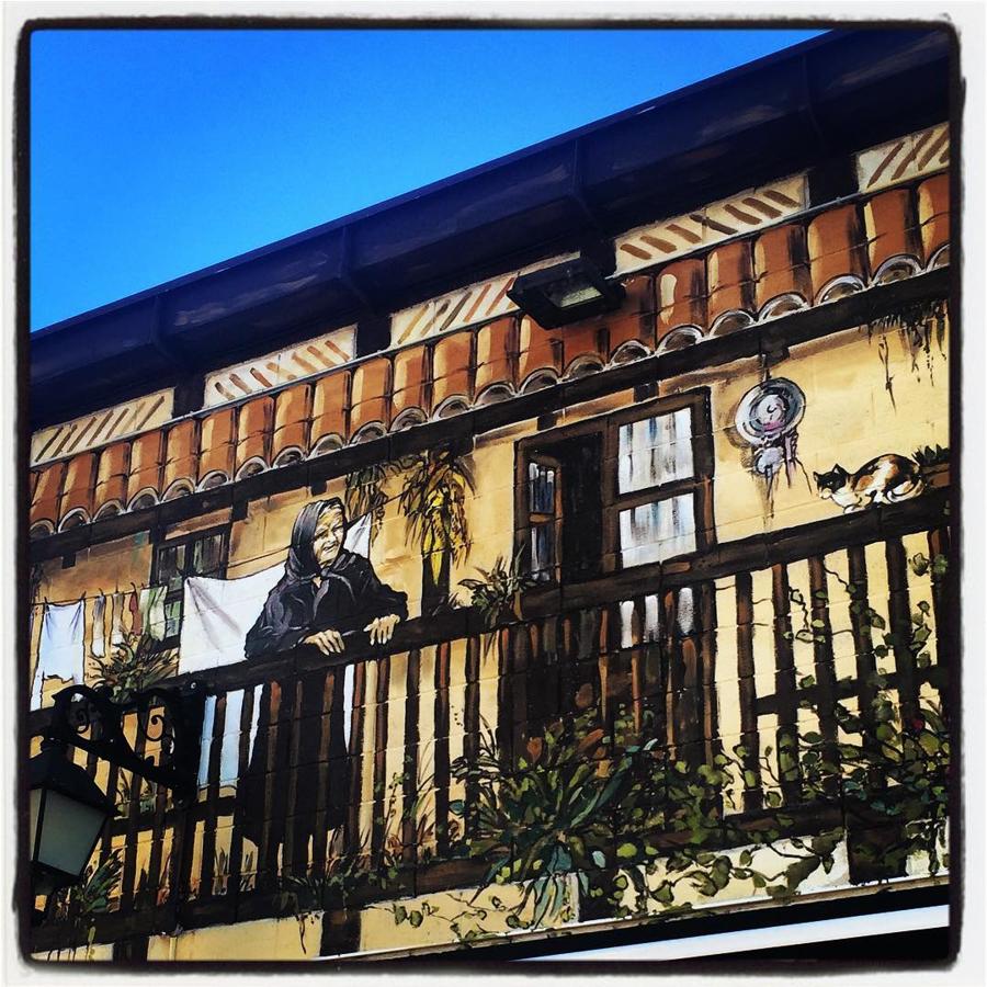 El restaurante Aurora Etxea, situado en la calle Las Trianas, luce en buena parte de su fachada un espectacular mural; obra de Pakiyo. En este fragmento, vemos a un gato doméstico sobre la balaustrada de madera.