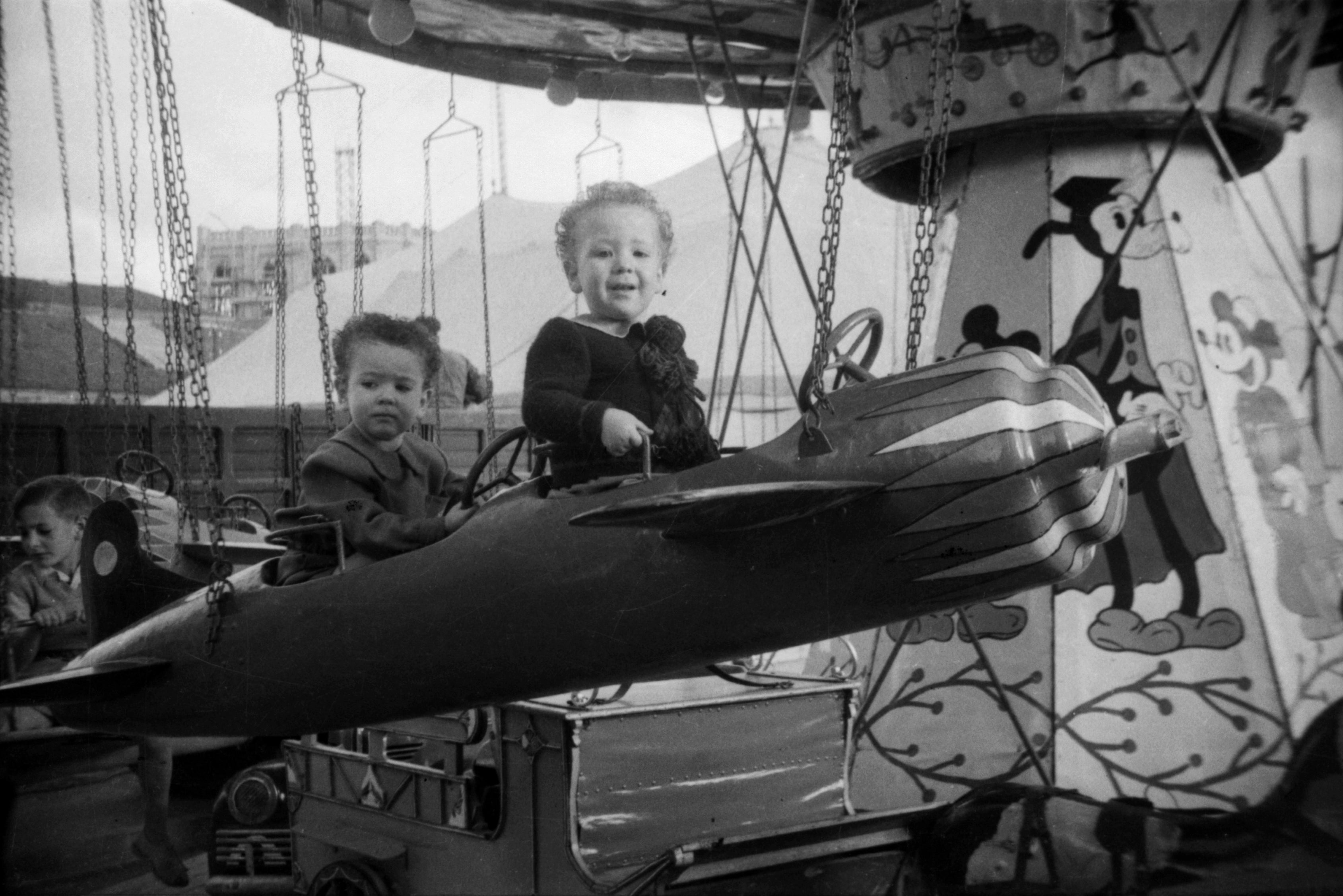 Retratos de dos niños de corta edad que montan en autos de choque y otras barracas y atracciones en el recinto ferial instalado entre las calles Becerro de Bengoa y Prado.