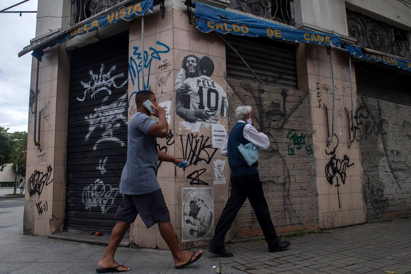 Hombres caminan frente a una pared de un bar cerrado con un grafitti del fallecido cantante jamaicano Bob Marley y el exfutbolista brasileño Pele abrazándose en el área del puerto en Río de Janeiro, Brasil, donde muchas tiendas permanecen cerradas debido a la pandemia de coronavirus, en diciembre de 2020.