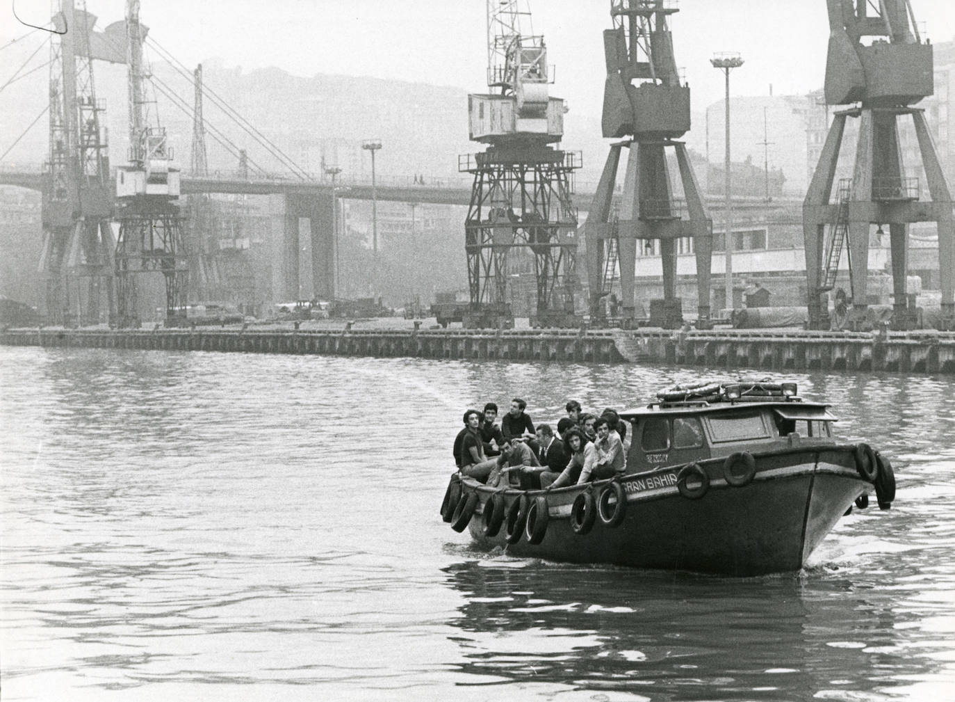 Un grupo de jóvenes viajan en un barco, con las fábricas de la Margen Izquierda de fondo. 