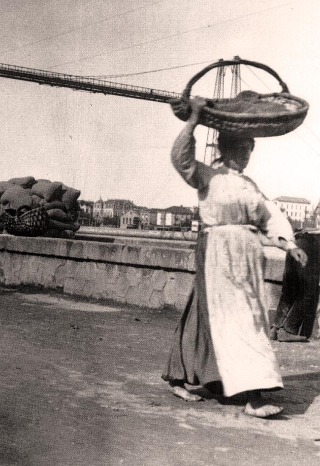 Una sardinera camina descalza por la ribera dejando tras de sí el Puente Colgante de Portugalete.