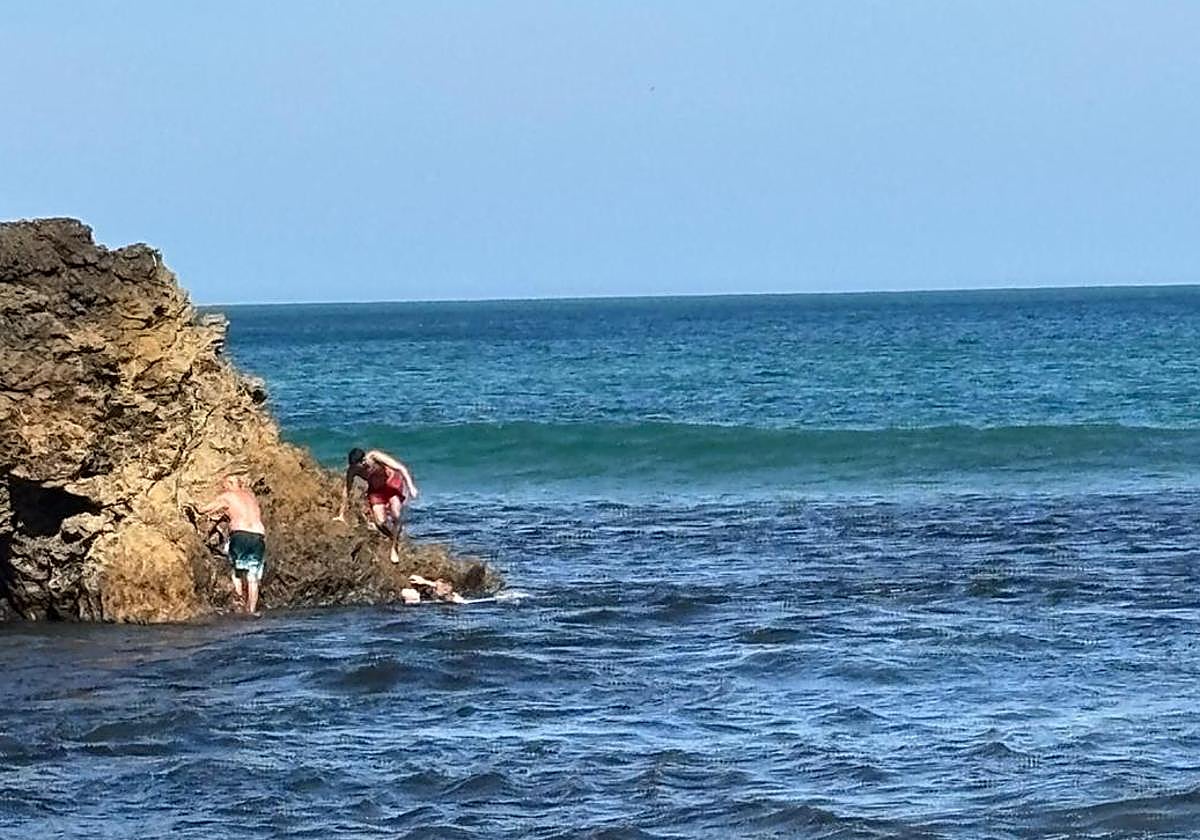 El hombre se agarra a la roca, mientras en el agua el ertzaintza sujeta a la mujer exhausta y el hombre del bañador rojo se acerca para ayudarlos.