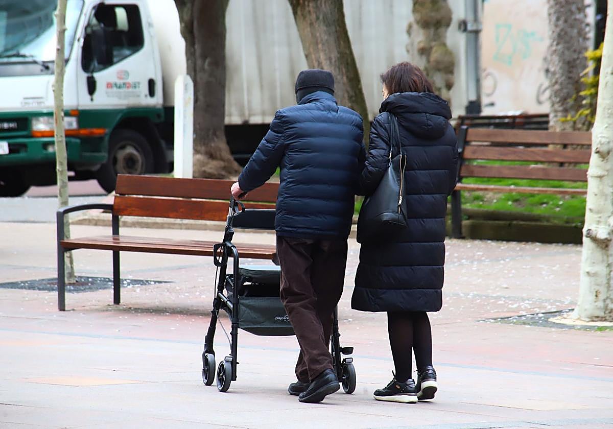 Una persona mayor camina ayudado por un andador y una mujer.