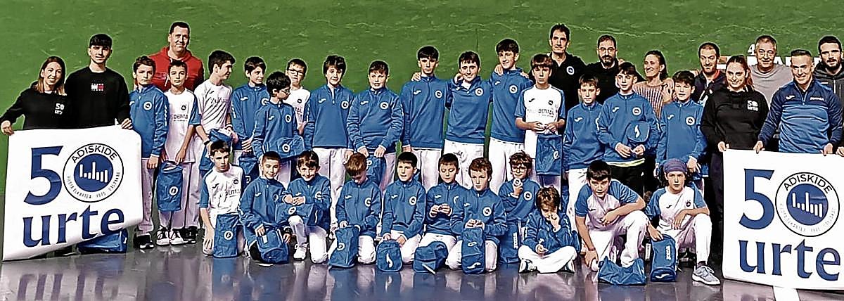 Las diferentes generaciones que integran el Adiskide posan en el frontón de Kurtzea, actual sede del club, con los carteles del aniversario.