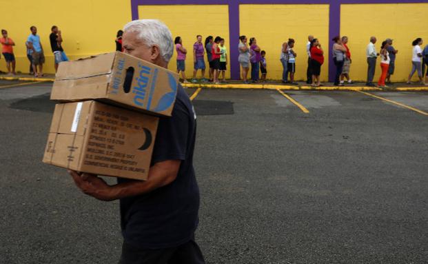 Un hombre camina con dos cajas de alimentos frente a una fila de personas damnificadas por el paso del huracán María. 