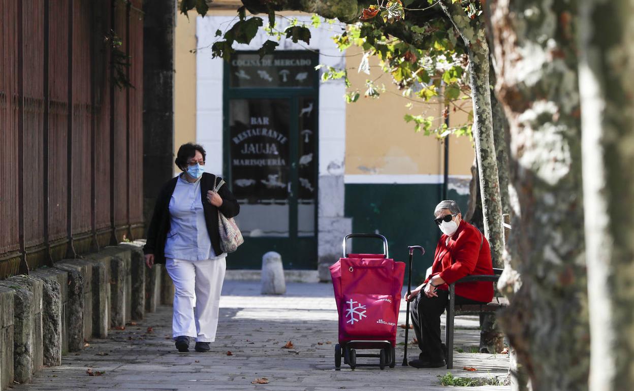 Una mujer camina por las calles de Santoña delante de otra que espera sentada con el carro de la compra, en el primer día de confinamiento.