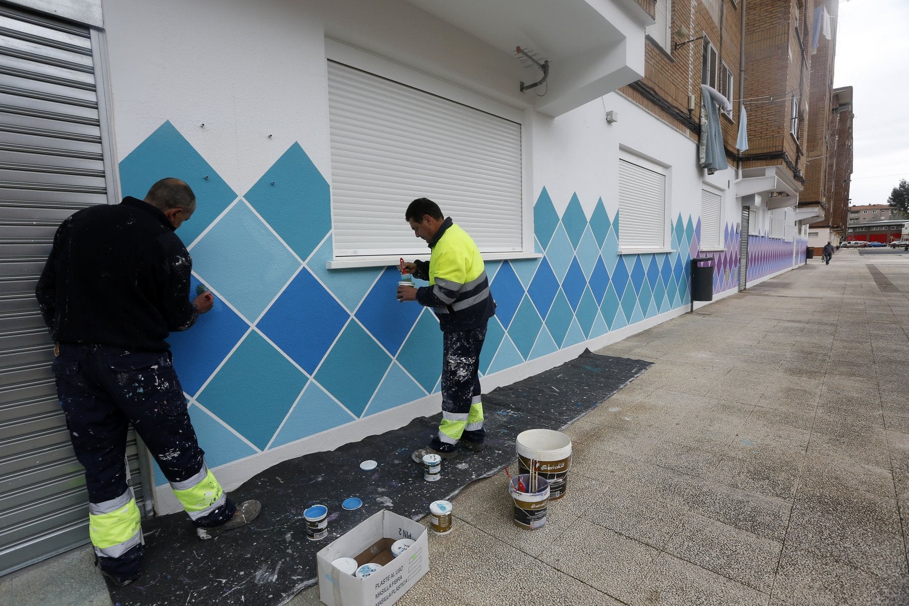 Dos trabajadores pintan un mural artístico en un local municipal de Nueva Ciudad.