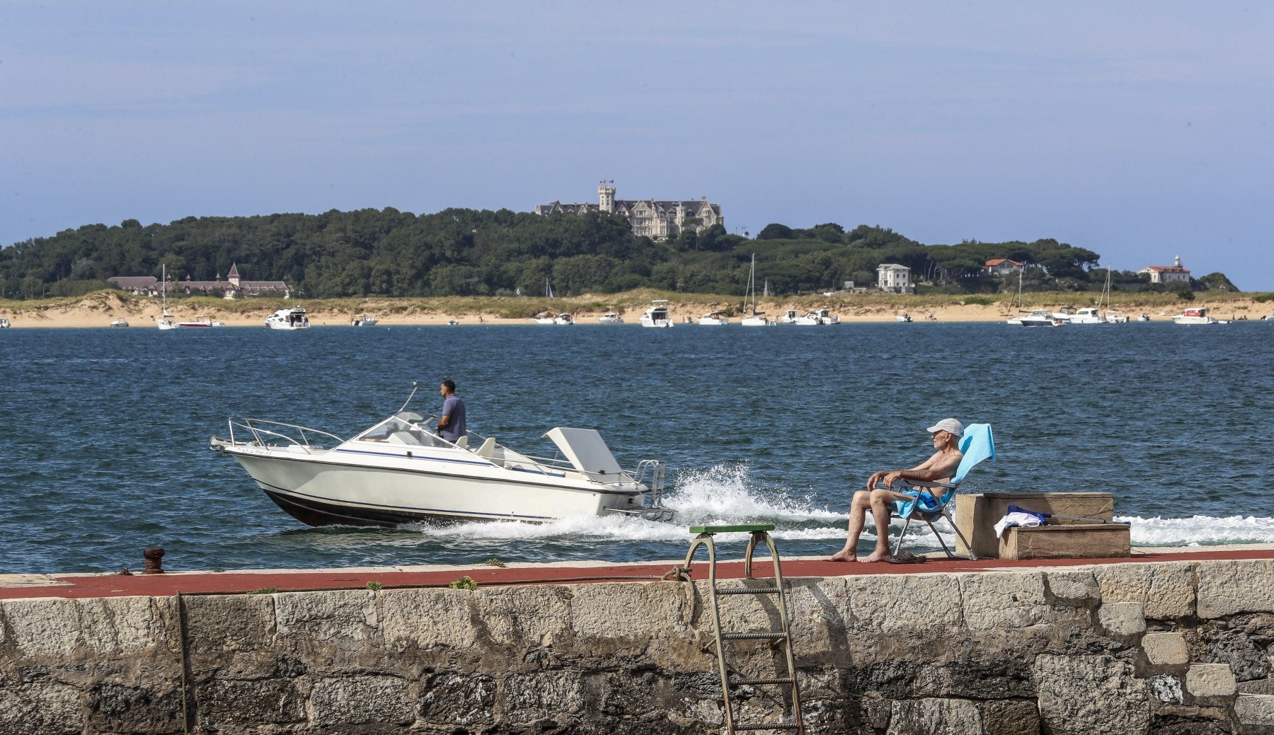 Un hombre toma el sol en el puerto de Pedreña mientras una motora abandona la localidad.