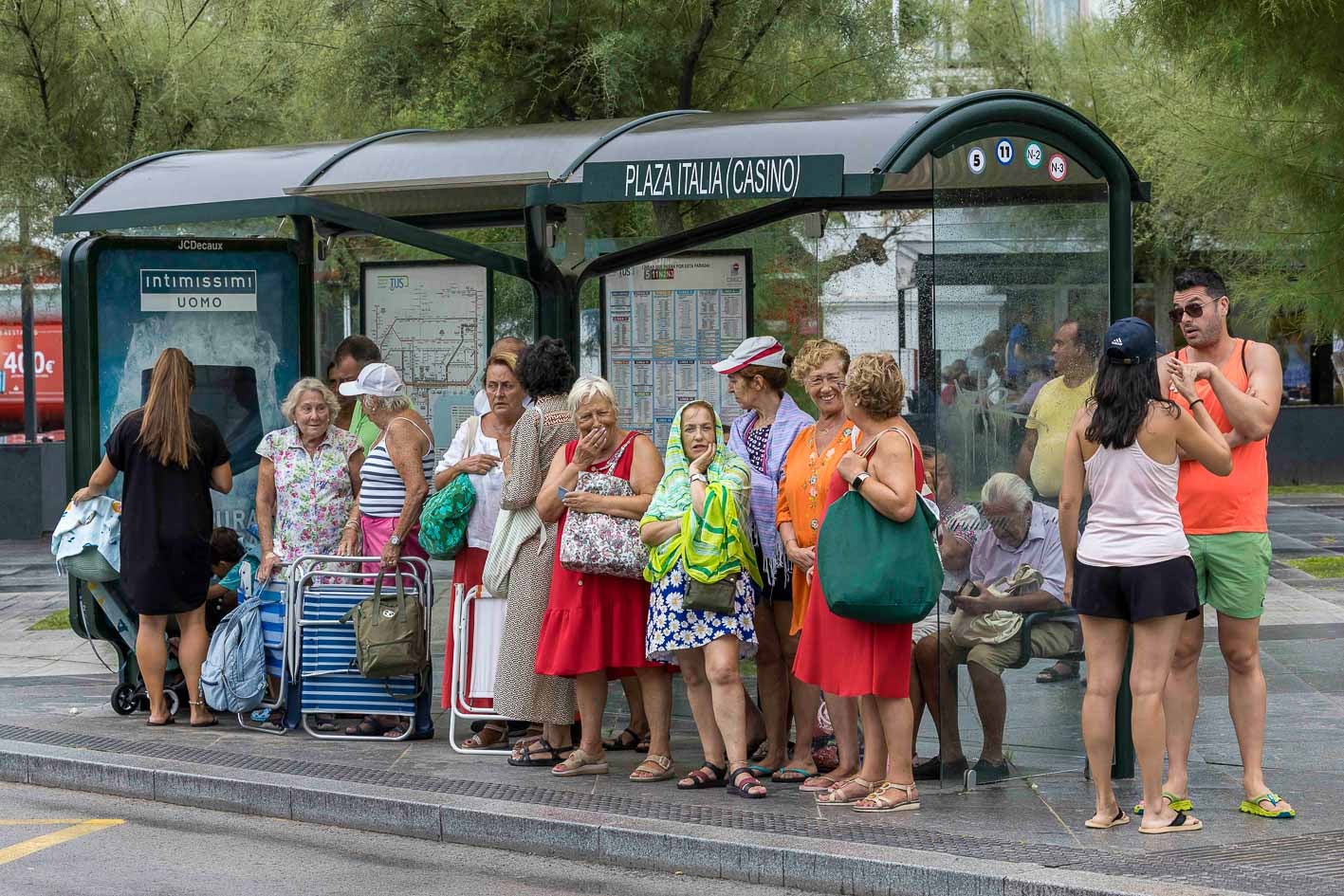 Varias personas esperan al autobús a la salida de la playa en una marquesina tras el chaparrón.