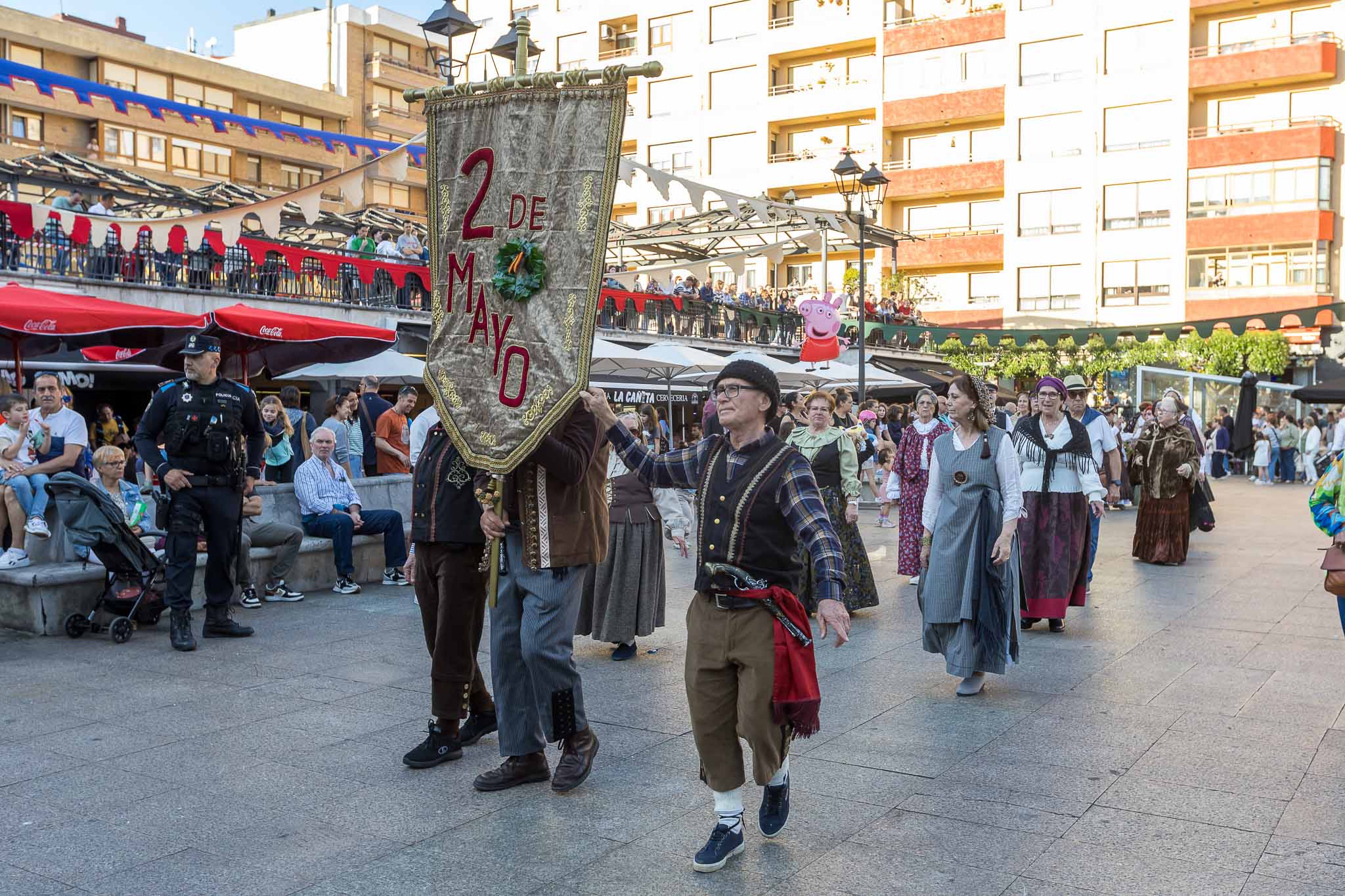 Unos hombres ataviados de época pasean por la plaza con un estandarte que anuncia la fiesta del Dos de Mayo. 