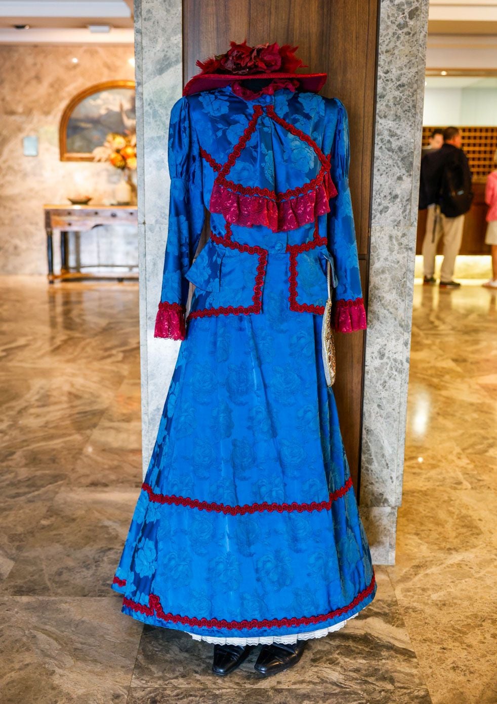 Traje azul y rojo de chaqueta y falda acampanada, adornado con puntillas de chantillí y sobrefaldilla forrada con el color de la puntilla. Se ultima el conjunto con un sombrero de tonalidades rojizas, un abanico pintado a mano y un bolso de pedrería.