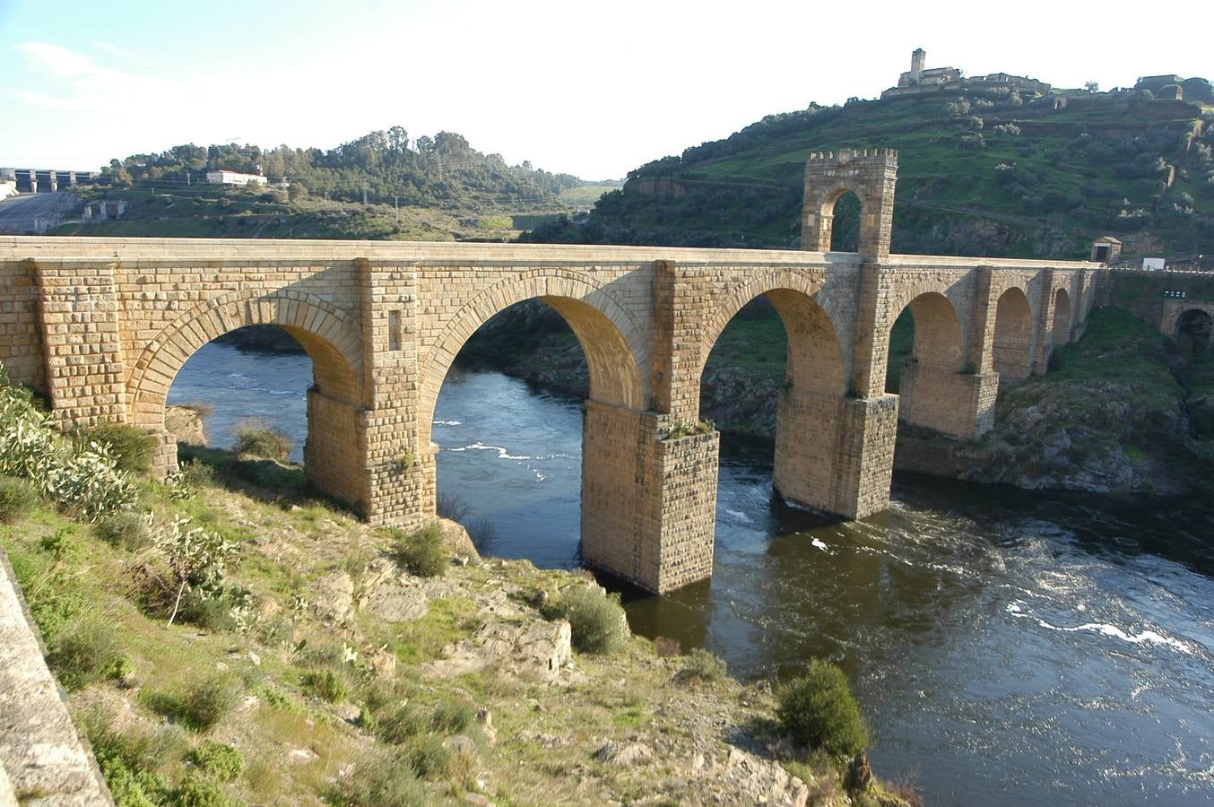 Alcántara (Cáceres). Cruza el río Tajo a su paso por la localidad de Alcántara. Es Monumento Nacional. Tiene seis arcos y, en la mitad, el llamado Arco de Trajano (a quien está dedicado el puente). A la entrada, un templete romano con esta inscripcion: «Este puente durará mientras dure el mundo».