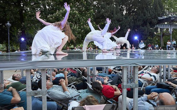Un grupo de espectadores recostados en hamacas bajo el escenario acristalado contempla a los bailarines de Imperdible en la plaza de Poniente. 