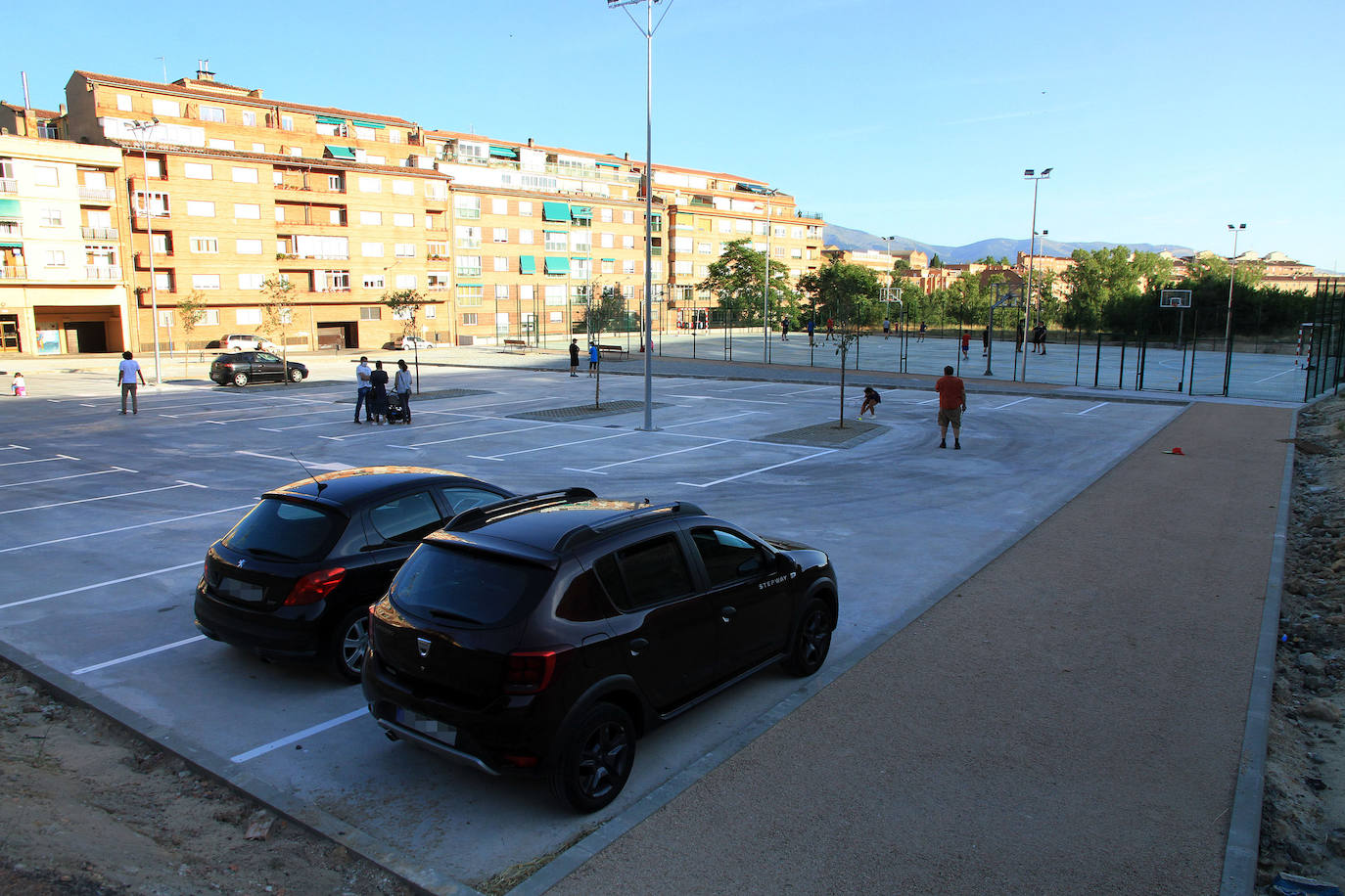 Dos coches estacionados en el nuevo aparcamiento, con la pista polideportiva al fondo.