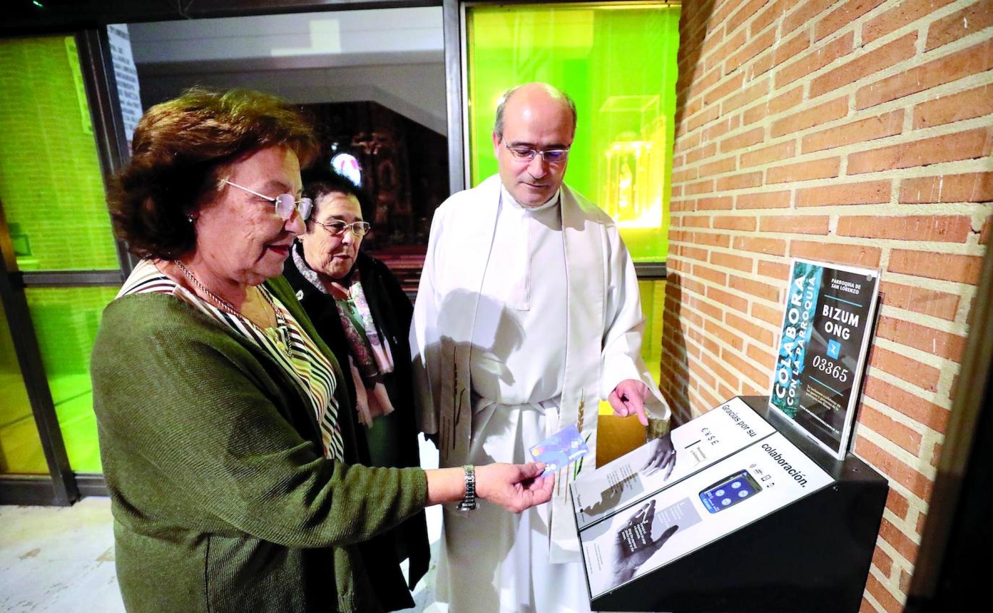Dos feligresas aplican la tecnología de pago con tarjeta en la máquina de limosna de la Iglesia de San Lorenzo, en Valladolid, ante su párroco, Jesús Álvaro Sancho. 