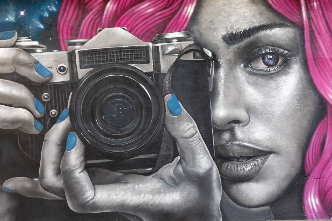 El rostro de una mujer con mirada penetrante, cabello rosa fucsia y uñas pintadas de azul sujetando una cámara que fotografía al espectador protagonizan este mural situado en las inmediaciones de la plaza Juan de Austria. Está dedicado a la fotografía del artista madrileño SHFIR.